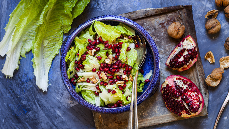 A leafy salad with nuts and pomegranate seeds sits next to leaves of lettuce, two segments of a pomegranate, and walnuts