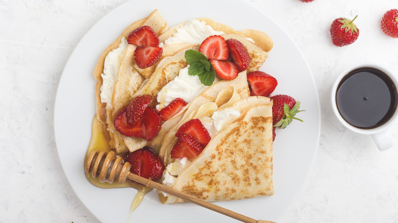 A stack of crepes is stuffed with strawberries and ricotta cream cheese on a white plate with a coffee cup to the right.