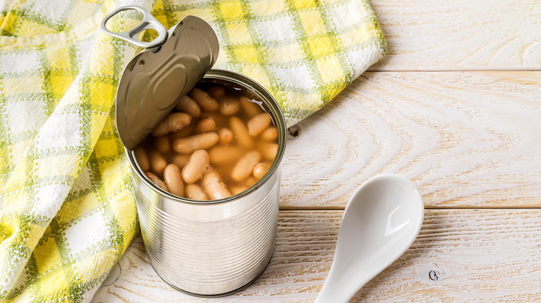 A can of pinto beans with the top peeled back, sitting against a yellow checkered cloth next to a white spoon