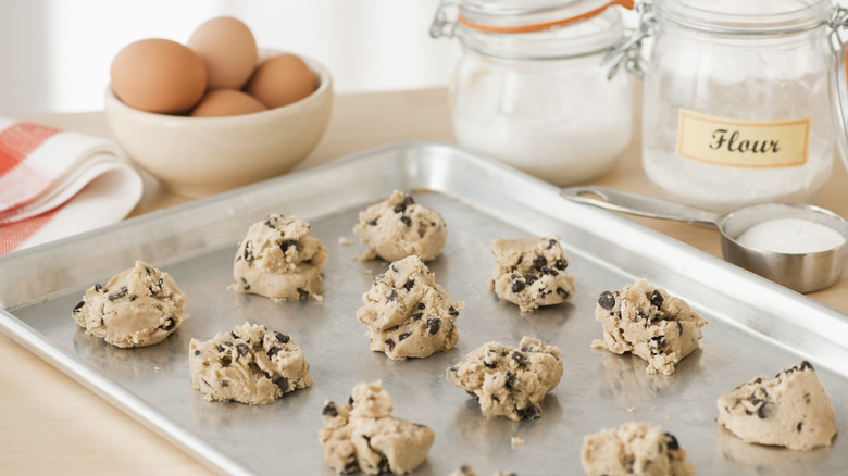 A baking sheet full of uncooked cookie dough balls sits on a counter next to a bowl of eggs and a container of flour