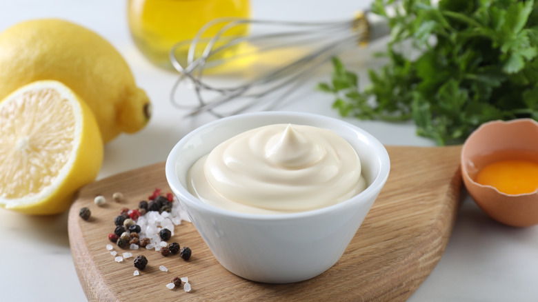 Mayonnaise in a white bowl surrounded by lemon, egg, and fresh herbs