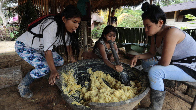 Three women chew and spit cornmeal into a large mortar and pestle to prepare chicha