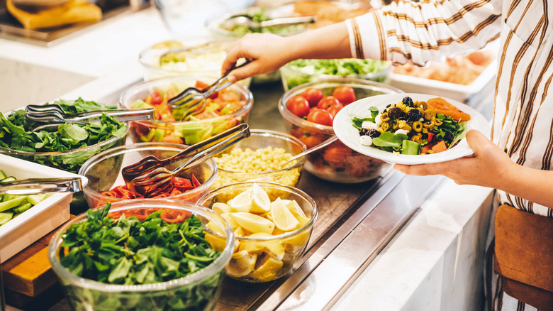 A woman uses metal tongs to grab fruit from a self-service salad bar