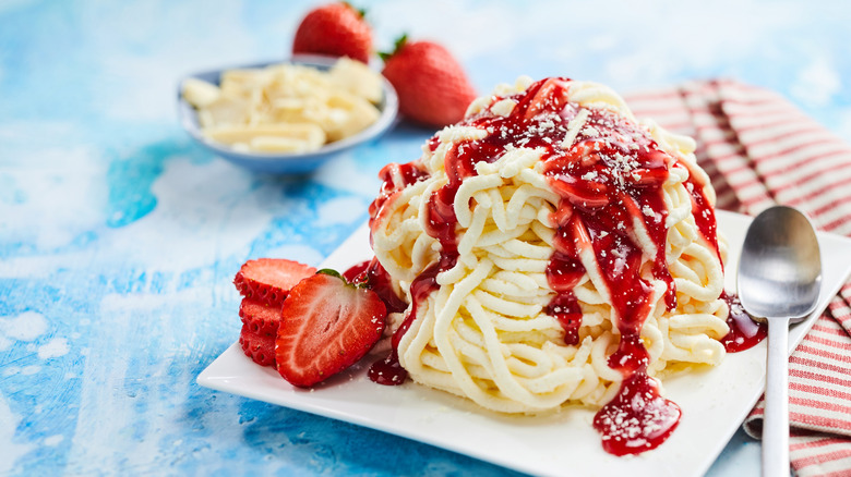 Vanilla ice cream that looks like spaghetti with red sauce on top and strawberries on the side and in background