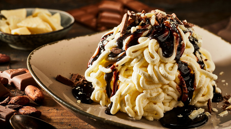 Vanilla ice cream that looks like spaghetti with brown chocolate sauce on a plate with nuts and chocolate on tabletop side