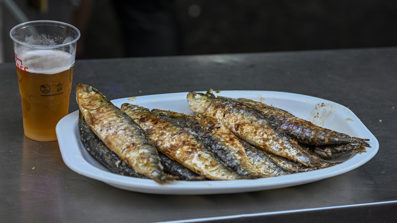 A plate of fried sardines sits next to a plastic cup of beer