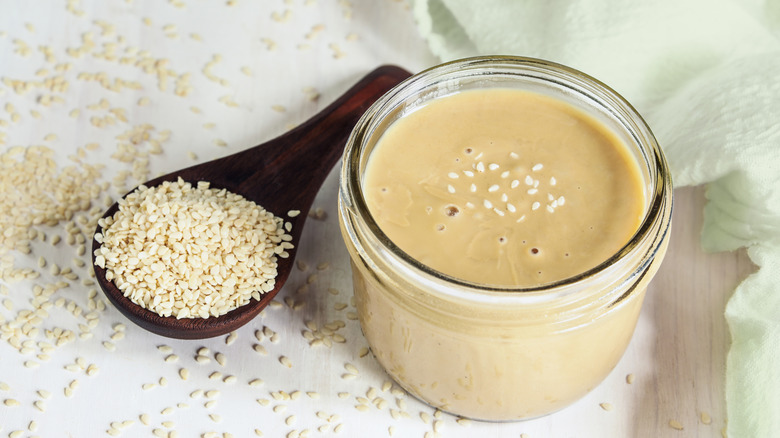 a wooden ladle of sesame seeds and a jar of tahini on a table covered in sesame seeds