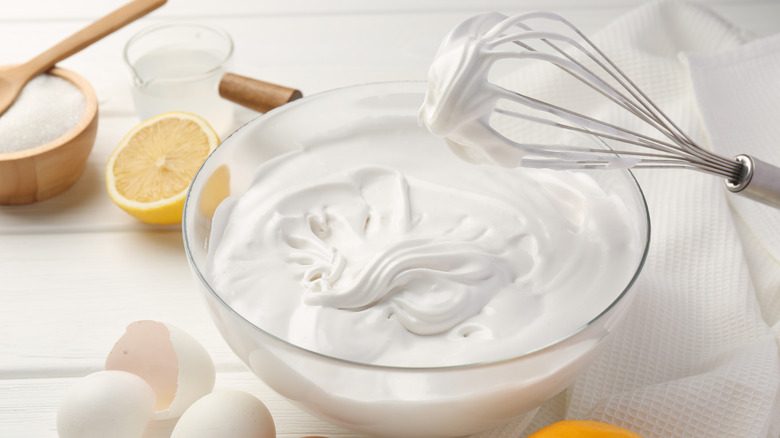 A large bowl of whipped meringue is surrounded by ingredients and a whisk