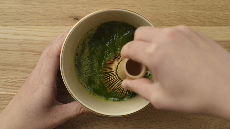 Hands whisking matcha with water in bowl using bamboo whisk