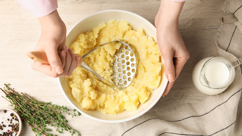 Woman's hands holding white bowl of potatoes while mashing them using a masher