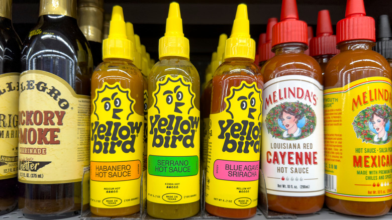 Three bottles of Yellowbird hot sauces on a shelf with bottles of other hot sauce on one side and a bottle of marinade on the other