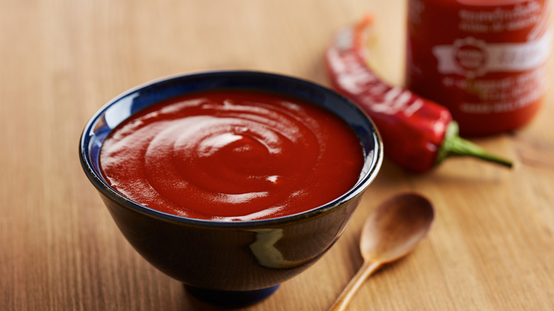 Bowl of red sriracha sauce on a wooden table with a spoon on the side and red jalapenos and a bottle of Huy Fong sriracha beside it