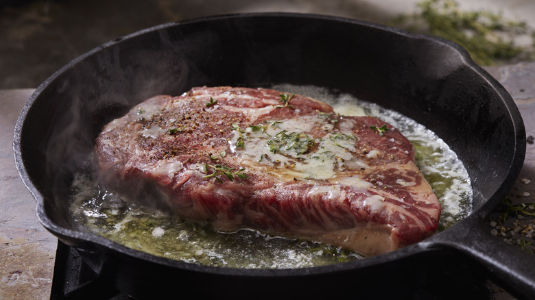 Raw rib eye steak being seared in a pan with butter and herbs