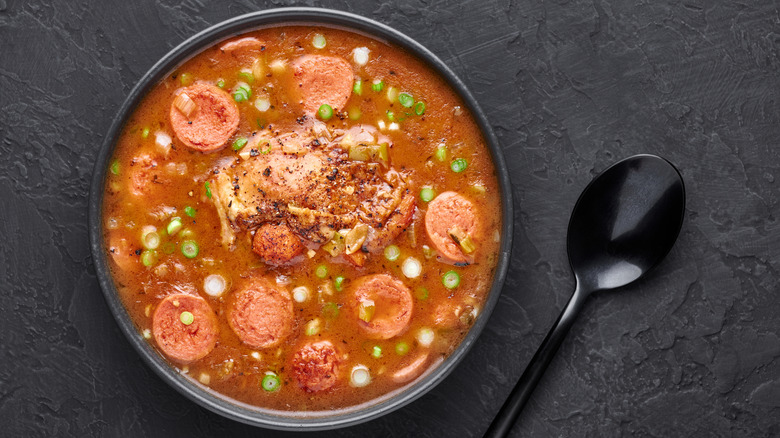 A bowl of gumbo made with dark roux sits on a gray surface next to a black metal spoon