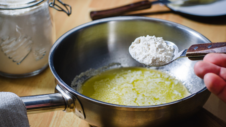 A hand pours a spoonful of flour into a pot of butter to make roux