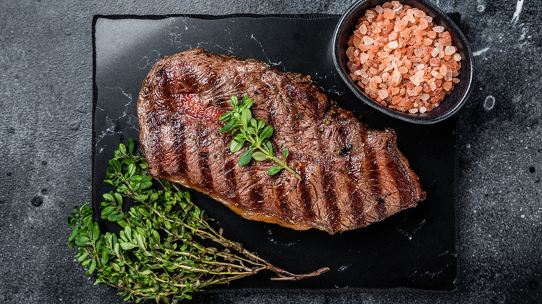 A beautifully grilled blade steak sits on a slab of black stone with herbs and salt