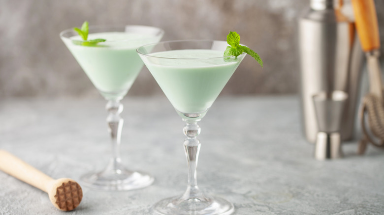 Two cocktail glasses with green grasshopper drinks, garnished with mint leaves, on a gray tabletop with a metal drink shaker and jigger in the background