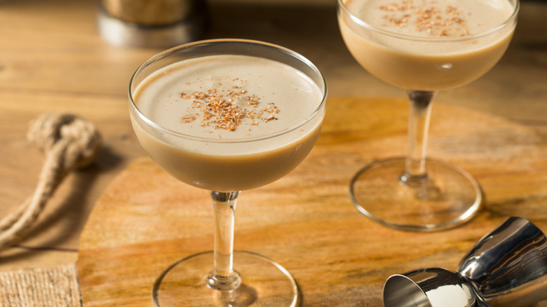 Two coupe glasses with light tan brandy alexander cocktails, on a wooden tabletop with a silver liquor jigger on its side next to them