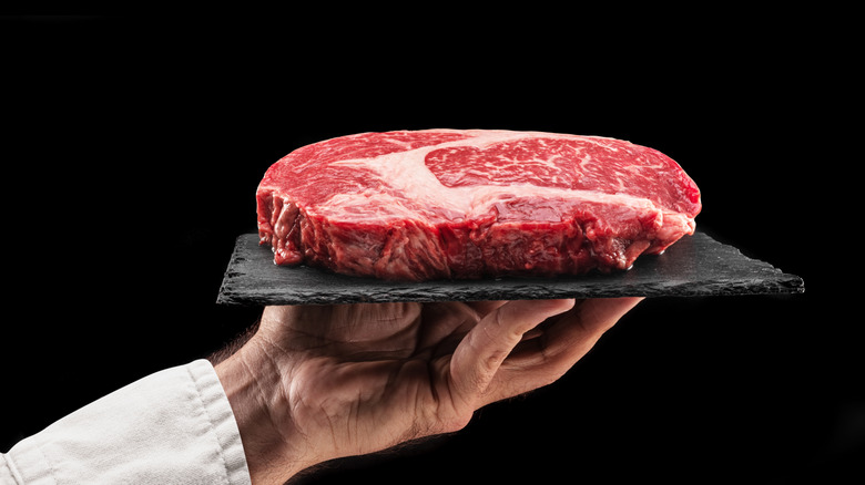 A hand holds up a slab of stone, on which rests a raw ribeye steak