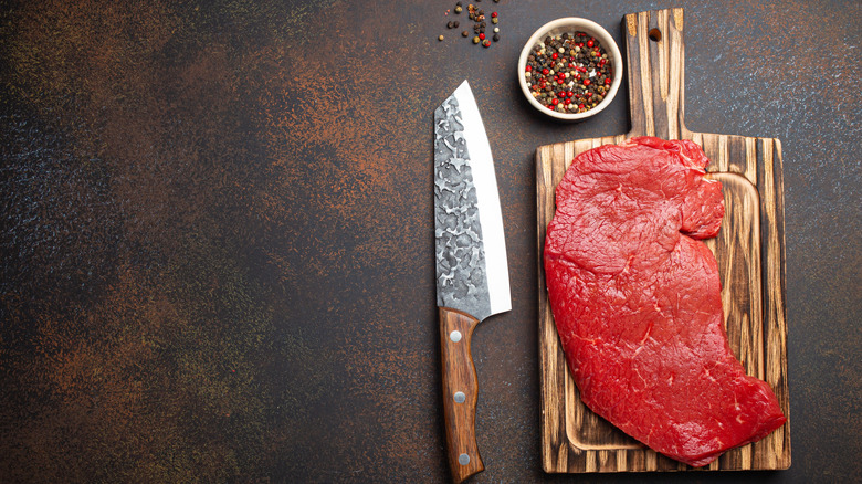 A brilliantly red piece of raw steak sits on a cutting board with a knife and seasonings near it