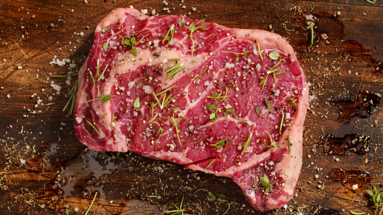 A raw ribeye steak sits on a cutting board, with herbs and spices on it