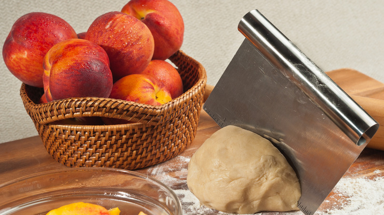 Bench scraper slicing dough next to bowl of peaches