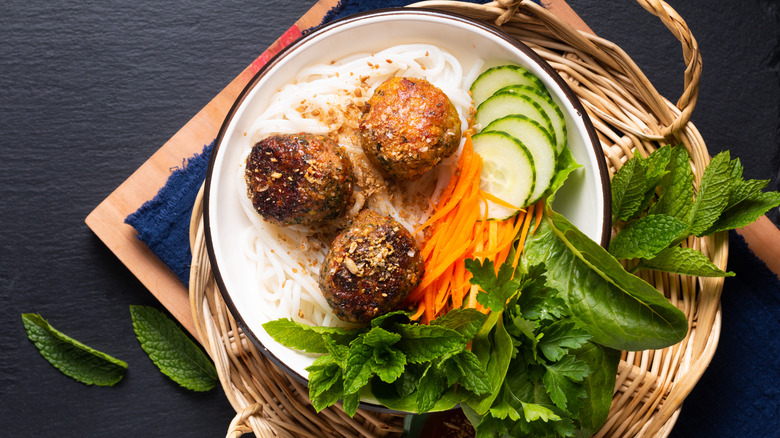 Three large meatballs sit on a bed of rice noodles, cucumbers, carrot shavings, and various herbs