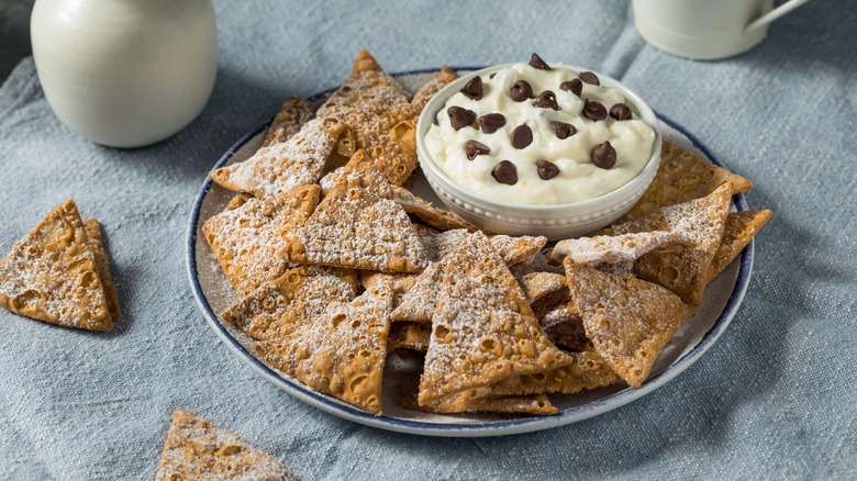 Small white bowl of white cannoli cream with chocolate chips on a larger white plate of golden brown cannoli shell chips dusted with white powdered sugar on a light blue cloth surface