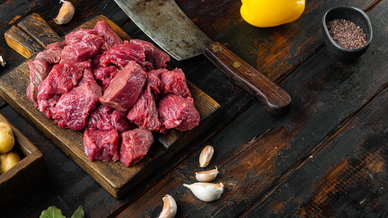 Diced raw chuck roast sits on a wooden cutting board next to a meat cleaver, with garlic cloves, a dish of seasonings, a yellow bell pepper, and potatoes nearby