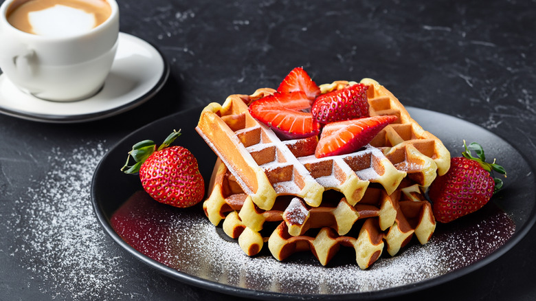 Stack of waffles on a black plate with strawberries and powdered sugar sprinkled on top and around. Cup of coffee with creamer on upper left corner all on a black marble tabletop