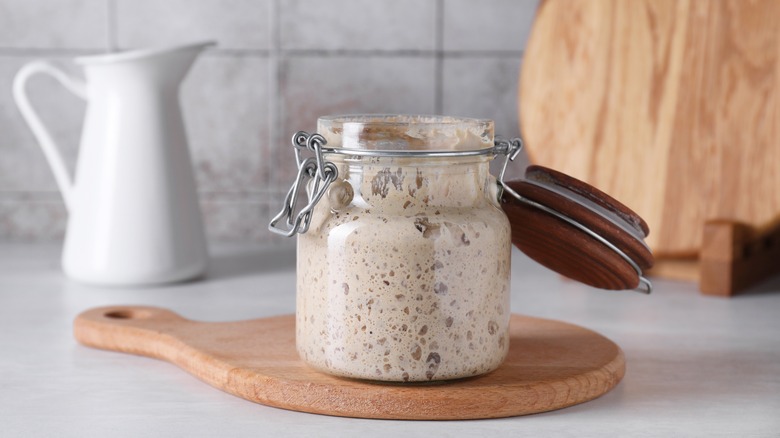 Beige sourdough starter in a clear glass jar with the lid hinged open on a wooden cutting board