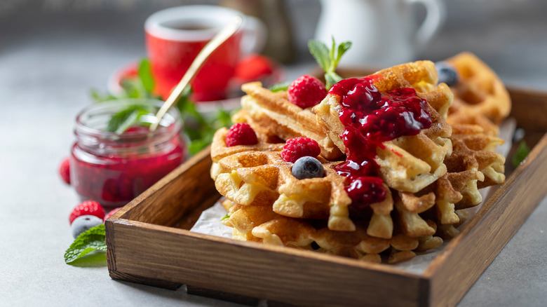 Small stack of sourdough waffles with red berry sauce on a wooden tray with a small jar of red sauce next to it