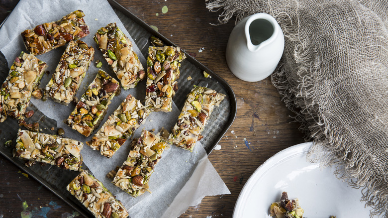 Homemade granola bars on a baking sheet with fruits and nuts on a wooden table, with a ceramic bottle next to the sheet.