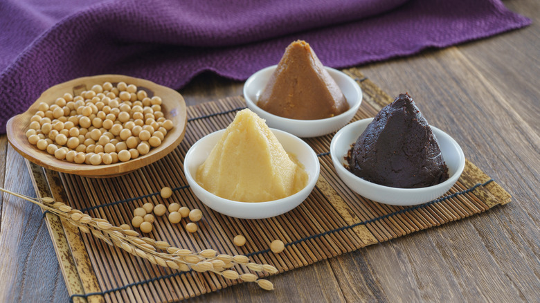 The three types of miso pastes, which are white miso, red miso, and yellow miso on a bamboo placemat and wooden table near a purple cloth.