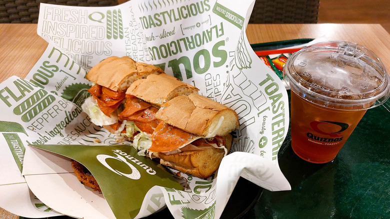 A Quizno's sandwich sits on a paper wrapper next to a Quizno's soft drink, both sitting on a plastic tray