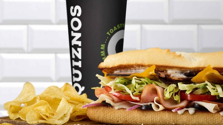 A Quiznos meal sits on a wooden slab, consisting of soda, sub, and potato chips