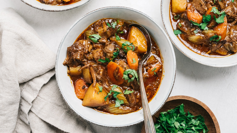 A close-up of three bowls of beef stew, one at the center, filled with tender beef, potatoes, and carrots and topped with chopped parsley, with wooden bowl of chopped parsley at the bottom right.