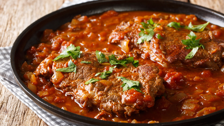 Two pieces of steak in chunky tomato sauce topped with parsley in a black pan on light wooden surface