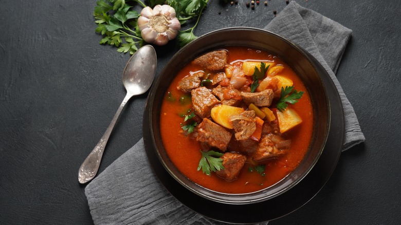 beef stew in a bowl on a table with spoon, cloth napkin and garlic bulb