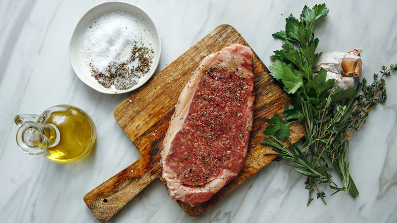 Seasoned raw steak on a cutting board surrounded by herbs, seasoning and olive oil