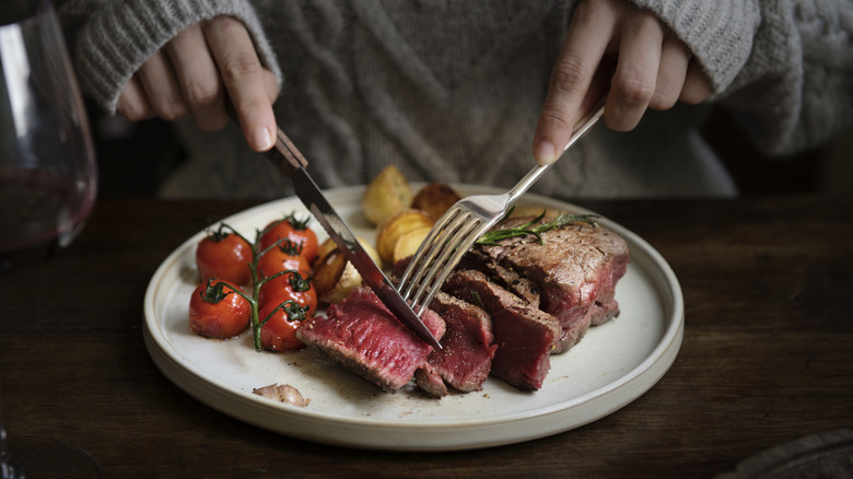 Woman uses a fork and knife to cut steak, roasted tomatoes, and potatoes