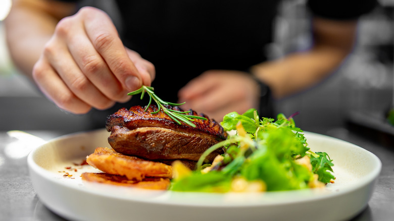 Restaurant chef adds fresh herbs to steak dish on white plate