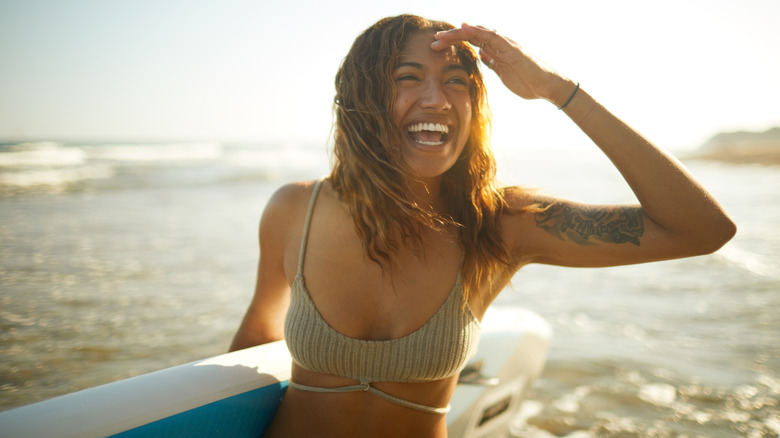 Woman carries surfboard near shallow ocean waves at sunset