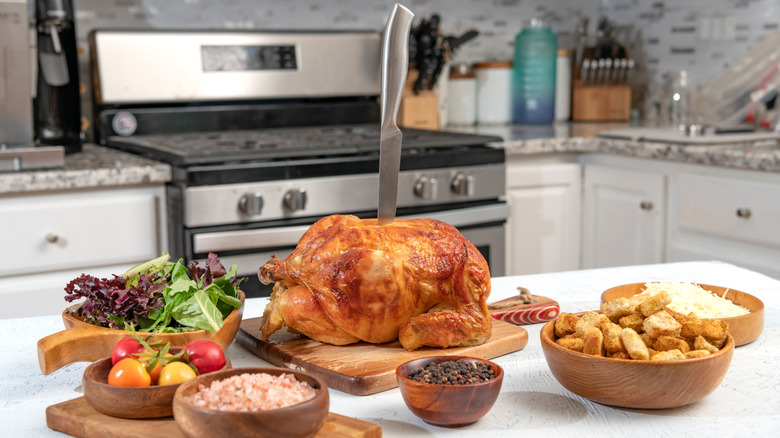 A counter with all the ingredients for chicken caesar salad and a knife sticking out of a rotisserie chicken