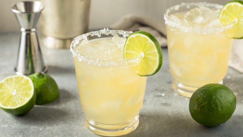two margaritas in tumbler glasses with jigger and limes around them