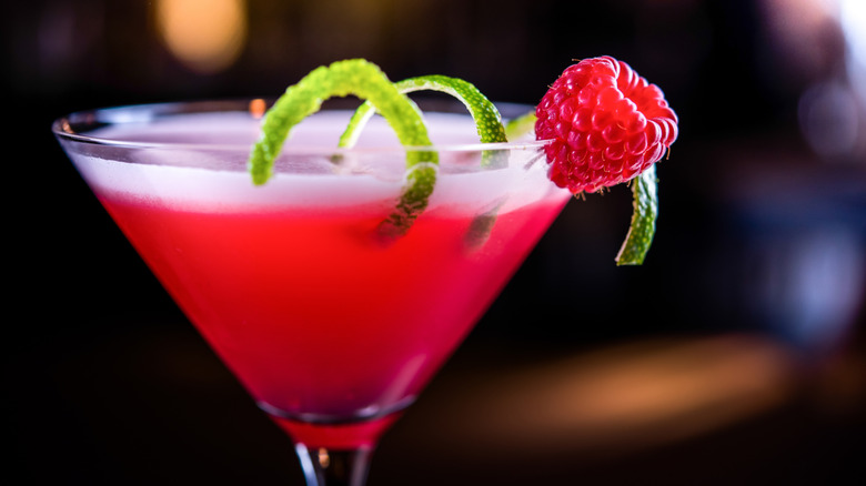 A reddish-pink cocktail in a martini glass with raspberry and lime peel garnish