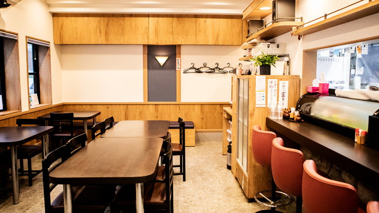 Interior of an izakaya with communal tables in the center, a bar with chairs to the right, and an open window into the kitchen. Smaller tables are along the wall on the left side of the room. Wood paneling and light walls are featured around the izakaya.