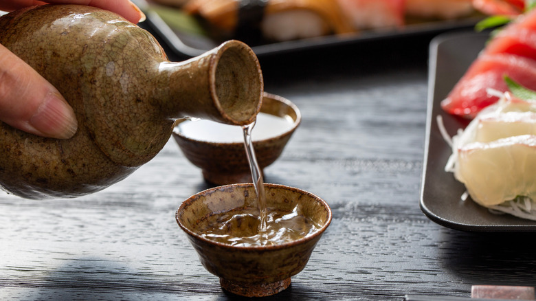 Hand pouring sake into small cup, with plated raw fish blurred to the right and in the background, all atop of a black, texturized table.