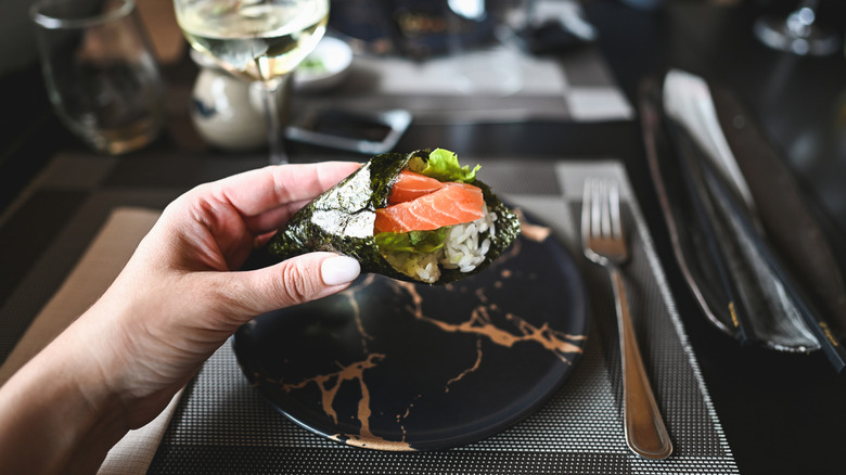 A hand holding a salmon temaki roll above a plate at a dining table.