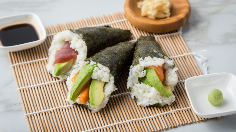 Three temaki, filled with tuna, salmon and avocado, on a bamboo rolling mat, with condiments in dishes.